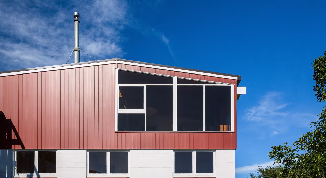 Exterior of Hillside Modern renovation, red coloursteel cladding and mid-century roofline under a wide blue sky, Christchurch.