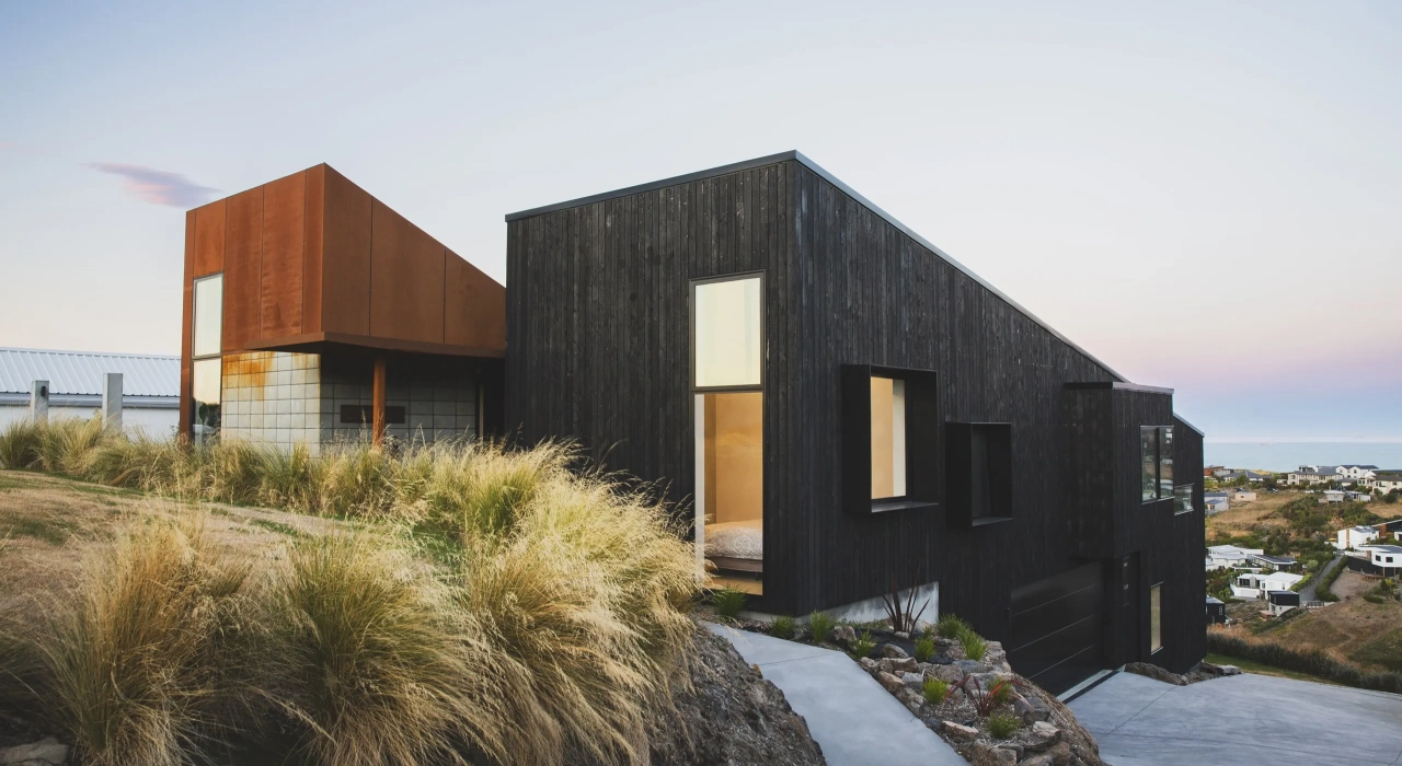 <img alt="Drayton exterior at dusk, charred larch and coretn steel volumes set among tussock grasses on the hillcrest." src="/images/s/i/t/e/-/site-architecture-drayton-drive8-70164273.webp?description" width="2560" height="1706" />